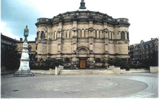 A wide expanse of circular patterned paving. There is a stone plinth with a large metal light on it to the left. The massive round neoclassical building behind has elaborate carved decorations, a large wooden door on the ground floor but no windows in its tall stone walls.