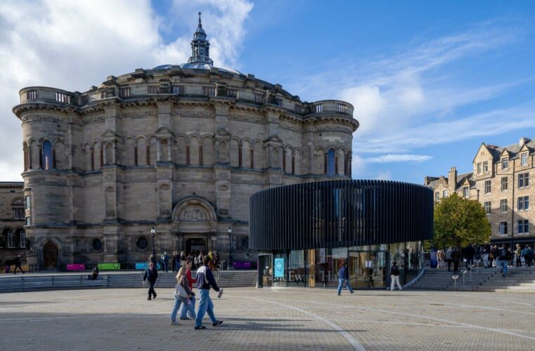 A wide circular paved area with people walking across it, deep steps around it and a low modern circular building with black slats around its top set into the steps. Behind that a huge round sandstone building with a domed roof topped by a spire. To the right sandstone tenements with regular windows.
