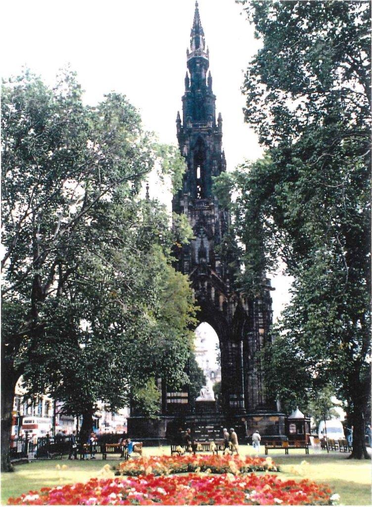 A formal garden with green lawn and two beds filled with mostly red flowers and tall green leaved trees on both sides. A huge, tall dark stone building at the back. It is pointed like a steeple and has ornately decorated stonework.