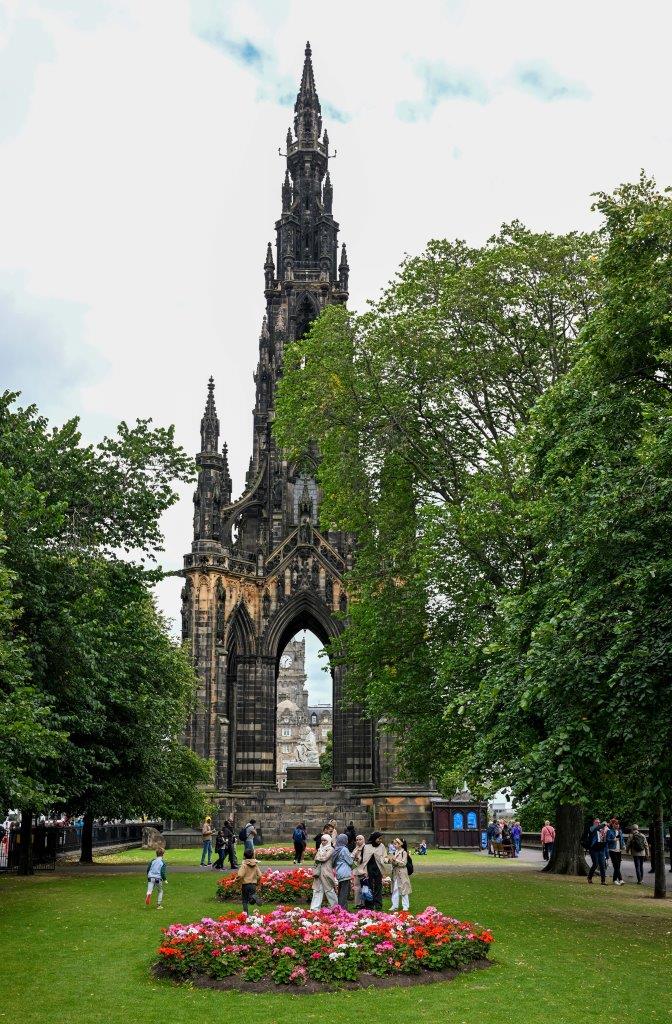 A public garden with people on the green grass lawn and tall green trees on both sides. There are circular beds of red, pink and white flowers. Behind a tall dark stone Gothic tower covered in ornate carved stone decorations rises high into the blue sky.