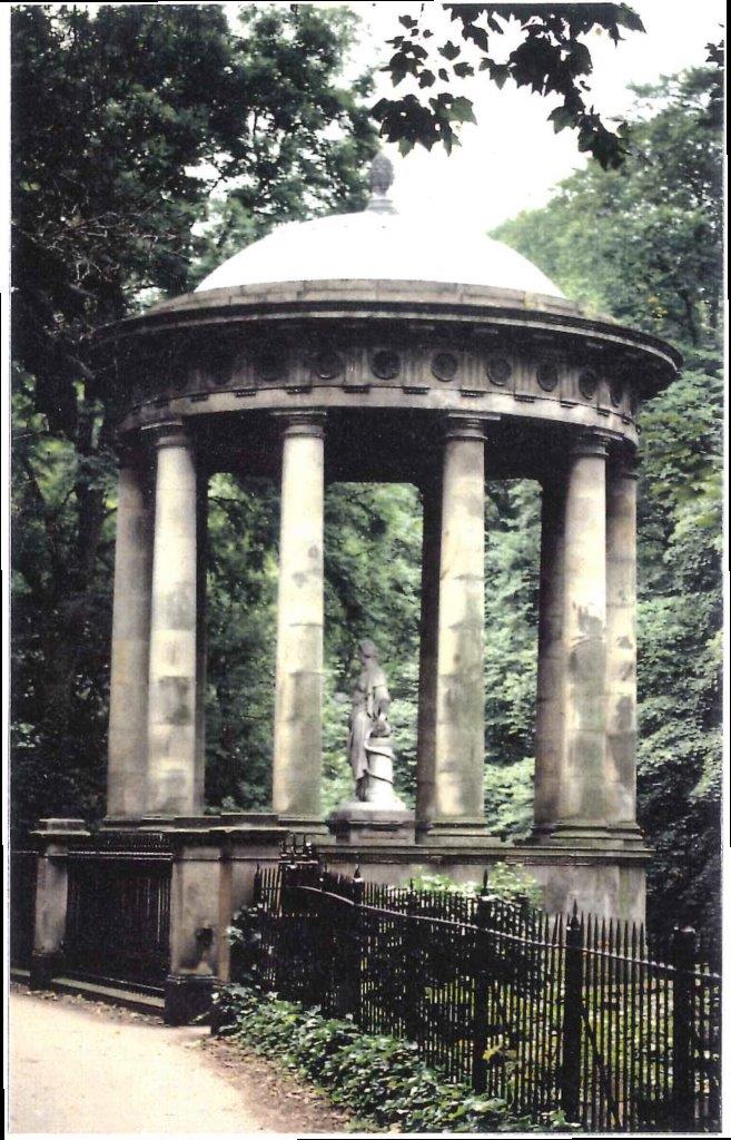A park with trees and lawns on either side of a central path that leads to a massive, tall stone column with a statue on top. The column is fluted and sits on a tall square plinth. The stone is darkened with age.