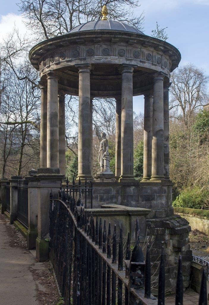 A park with trees and lawns on either side of a central path with many people walking there, it leads to a massive, tall stone column with a statue on top. The column is fluted and sits on a tall square plinth.