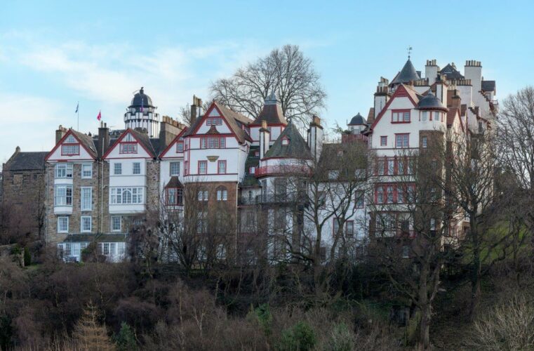 A group of white buildings with steep red tiles roofs on the brow of a hill. Bare trees in front and behind them and blue sky above.