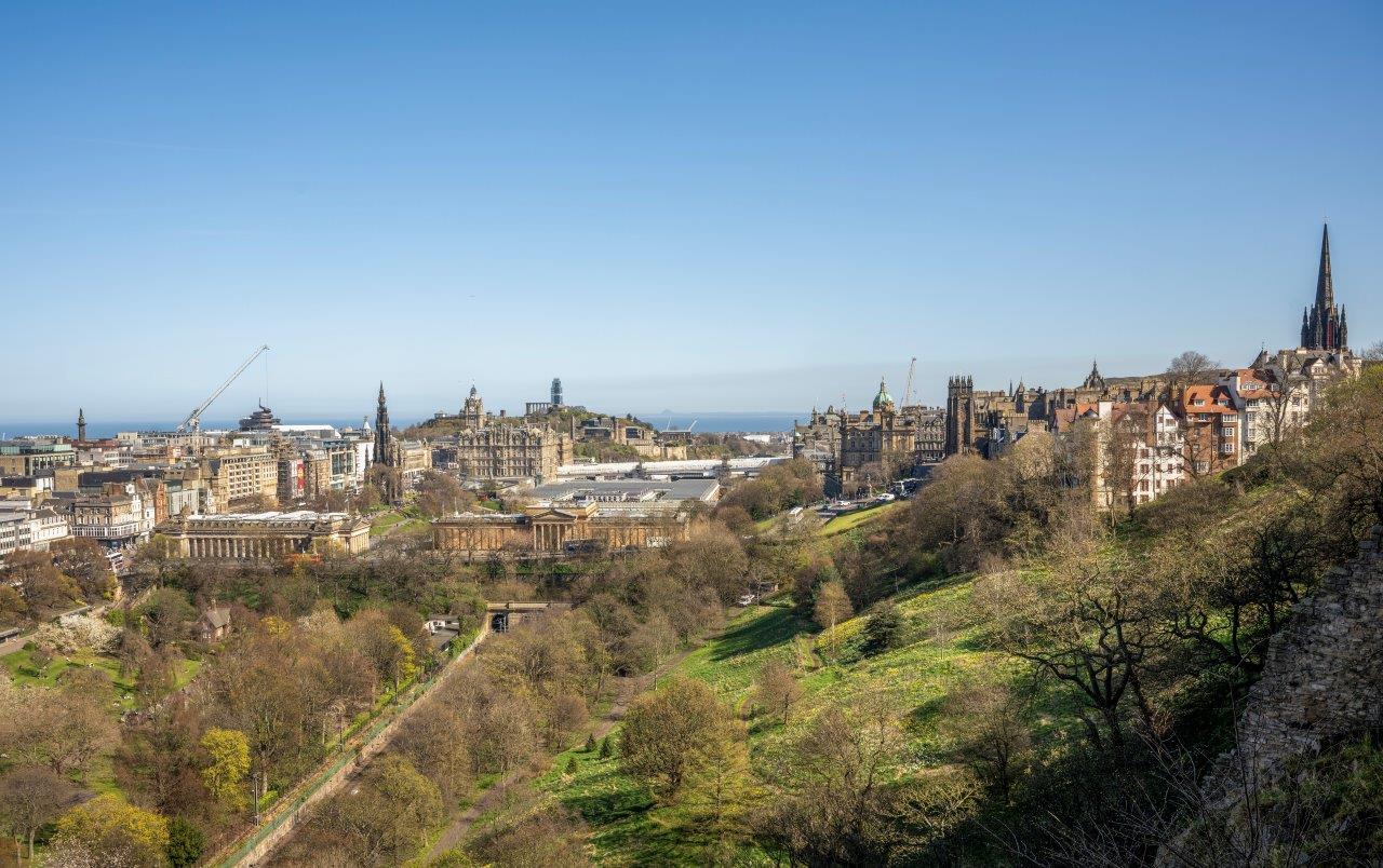 A large public gardens with green grass and trees at the bottom of a hill. Beyond lies a city with old and new buildings and the sky blue above.