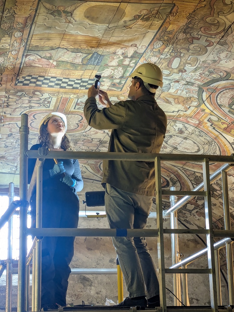 Two individuals wearing hard hats stand on scaffolding, inspecting an ornate, painted ceiling. One person holds a tool or device up toward the ceiling, which features intricate artwork with historical or religious motifs.