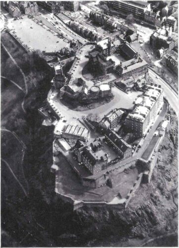 Edinburgh castle from above in black and white. On a high rocky hilltop many stone buildings of different types surrounded by a high wall. A large paved area with parked cars in front of it and the city surrounding it below.