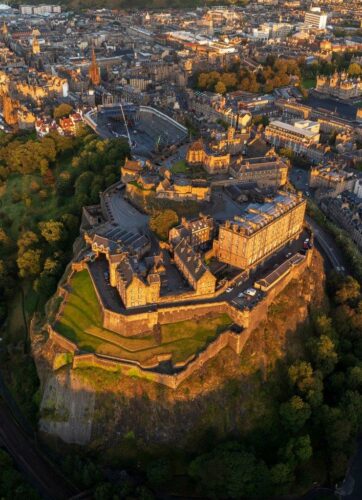 Edinburgh castle from above in colour. On a high rocky hilltop many stone buildings of different types surrounded by a high wall all glowing in the warm sunlight that falls upon them. In front of the castle a large grandstand of seats and below it the city streets.