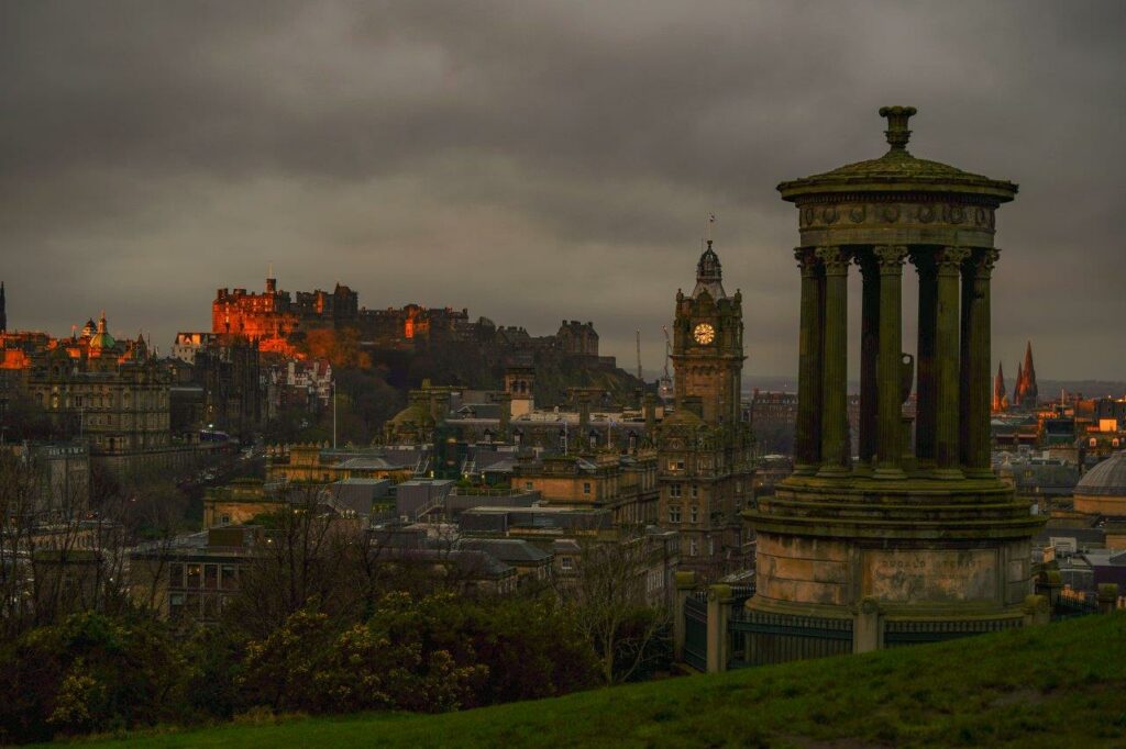 A round stone neoclassical temple building on a hill with a city roofscape below and a stone castle on a hill in the distance it’s walls red from the setting sun. The sky is dark and cloudy above.