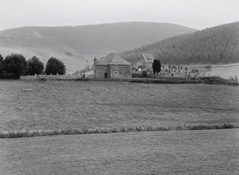 A small church sits in the middle of a rural landscape, with hills in the background and a field in front.