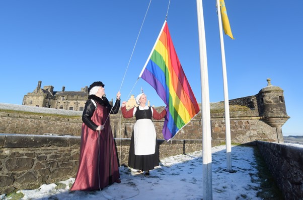 Two women dressed in historical outfits raise a rainbow flag on a Castle. There is snow on the floor.