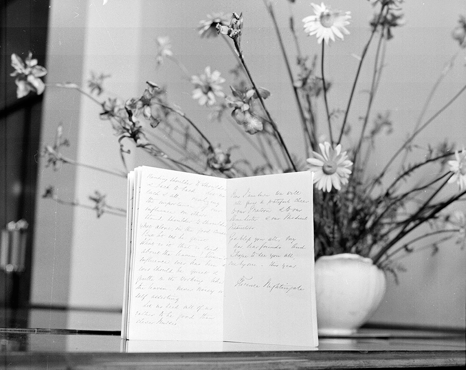 black and white image of folded paper covered in elegant handwriting, standing on a surface in front of a vase of tall daisies