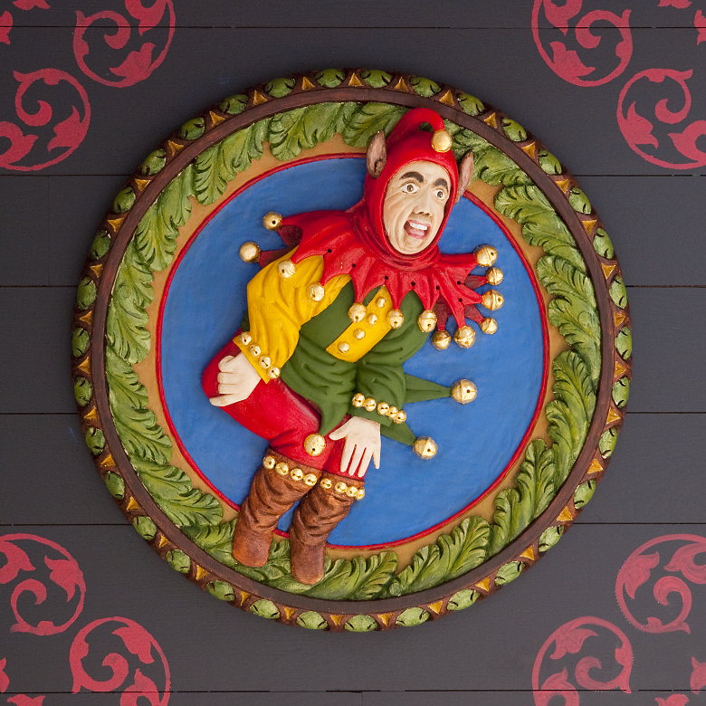 Colorful carved oak ceiling panel known as a Stirling Head, depicting a jester in vibrant costume with red, yellow, and green sections, adorned with gold bells. The figure is set against a bright blue circular background framed by a decorative green and gold border, with additional red scrollwork patterns on the surrounding surface.