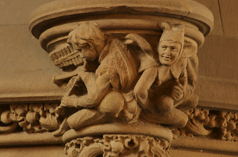 Detailed stone carving on a column capital depicting two crouched figures in intricate medieval-style attire. One figure is playing a pipe. The other figure is a jester, wearing a distinctive jesters hat and ruff. The carving includes ornate decorative patterns along the base and exhibits fine craftsmanship with rich texture and depth.