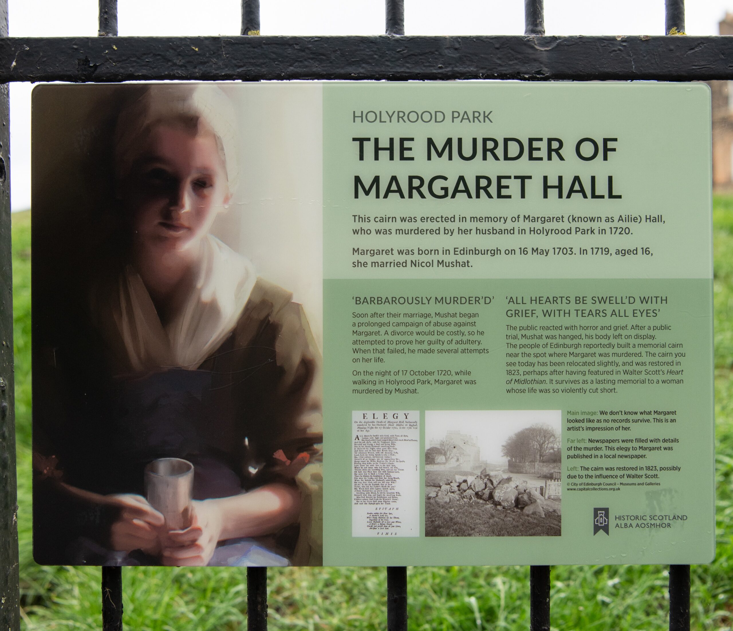 Information board at Holyrood Park titled “The Murder of Margaret Hall.” The panel includes historical details about Margaret Hall, who was murdered by her husband in 1720, along with excerpts of text, an archive photograph, and a depiction of a person in period clothing holding a metal cup. The board is mounted on black railings with grass visible in the background.