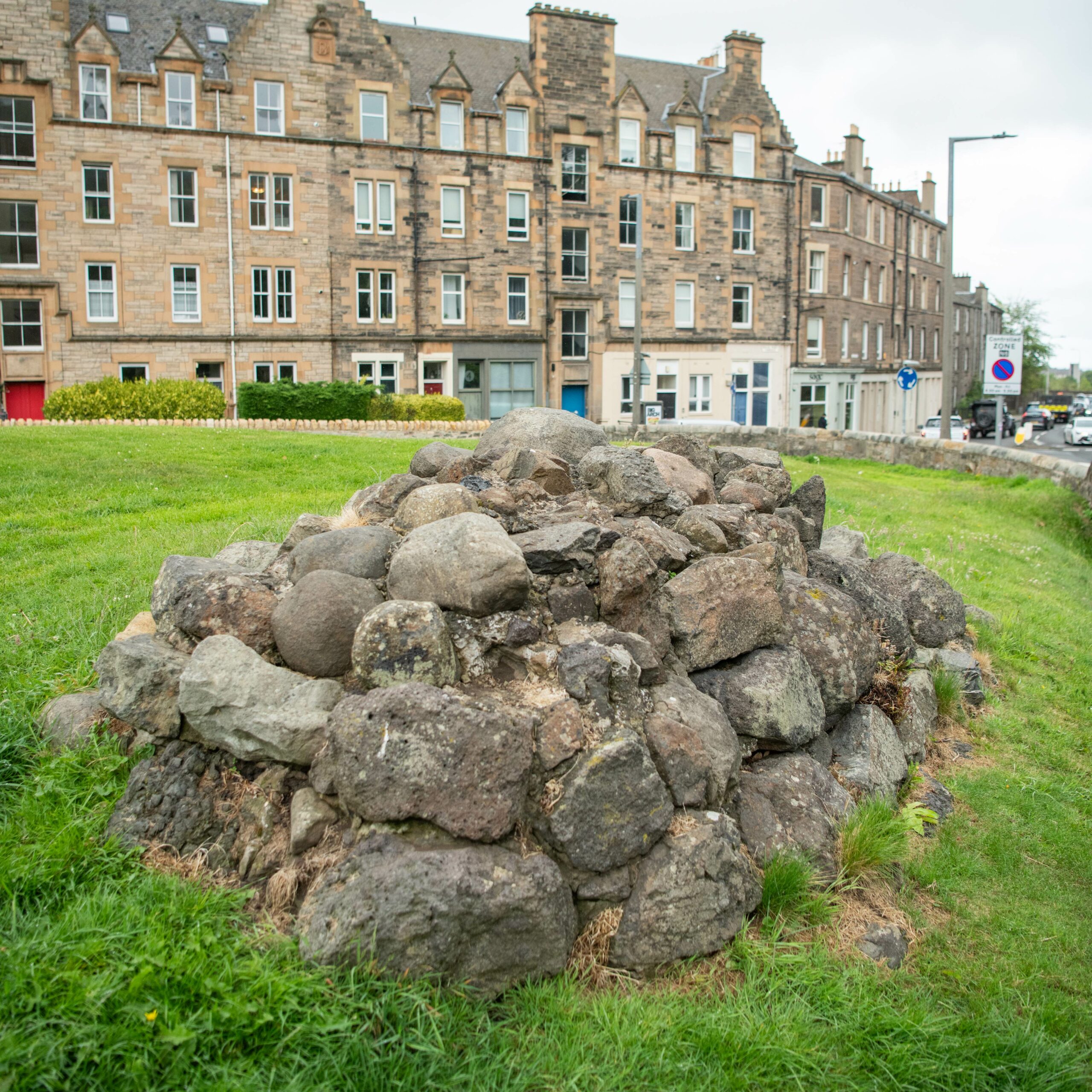 A cairn made of large, irregular stones sits on a grassy area near a roadside. In the background, there is a row of tall, historic stone tenement buildings with multiple windows and chimneys. A street with parked cars and a traffic sign is visible to the right.