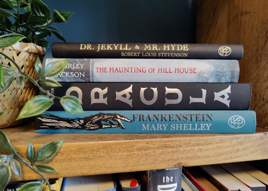 Selection of horror novels on a wooden shelf with a plant next to them.