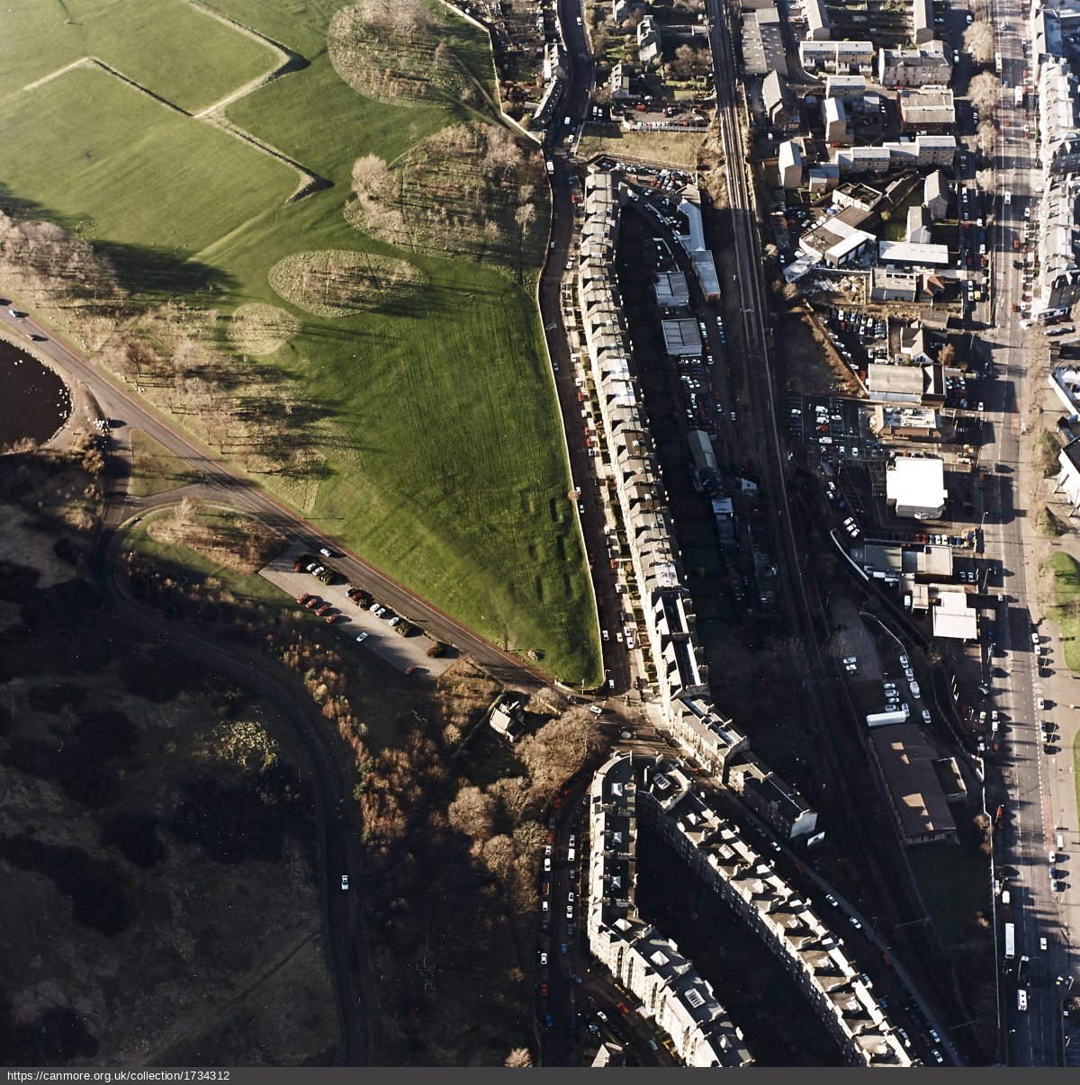 Oblique aerial view of Holyrood Park in Edinburgh, showing a large green open space with scattered trees on the left and a dense urban area on the right. A curved road runs along the edge of the park, and rows of terraced buildings are visible near the railway tracks.