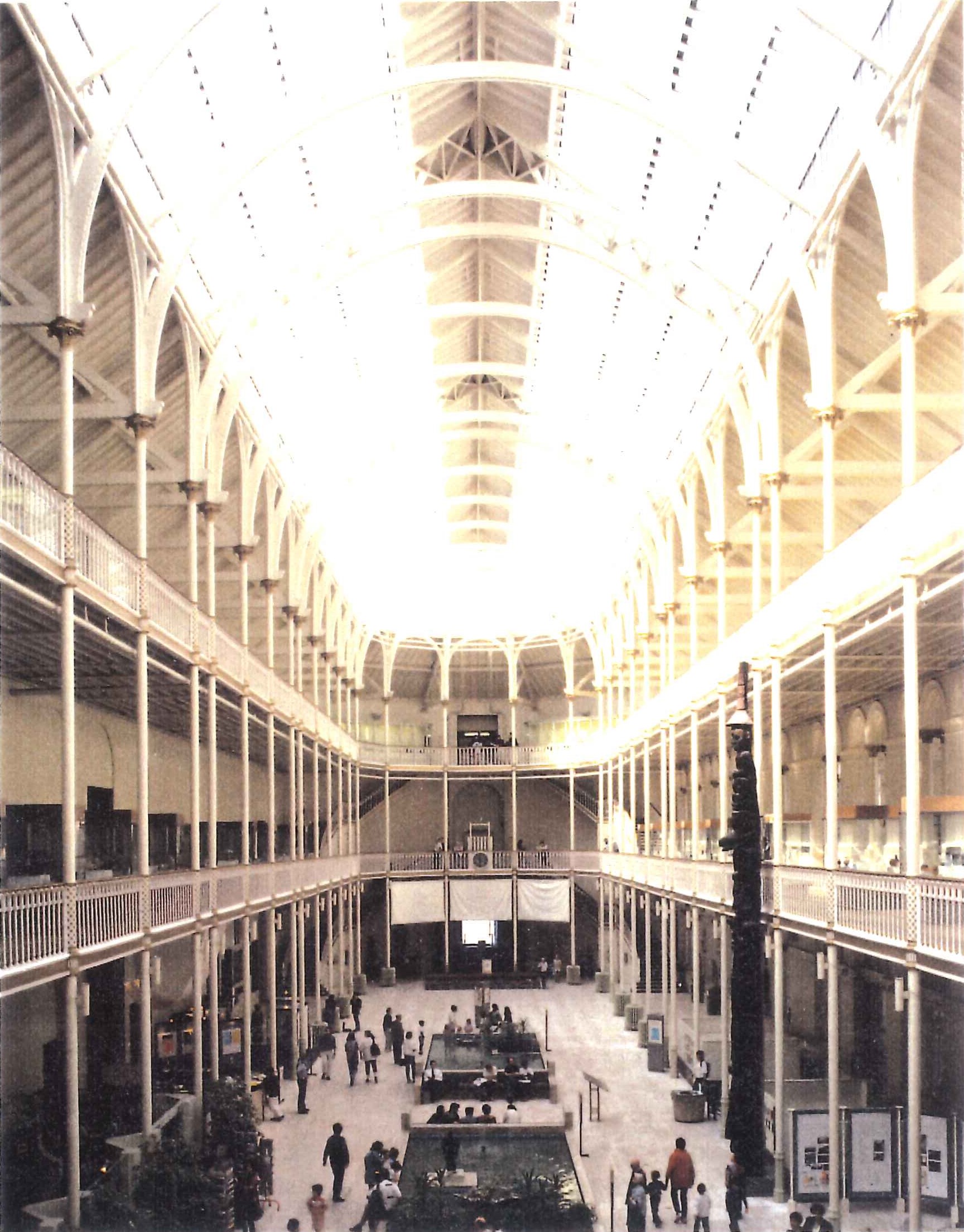 A large three storey room with two deep galleries held up by decorative white cast iron columns around three walls. Light floods from above, the ceiling is high and supported by white iron framework. The floor is pale marble busy with people, two large rectangular ponds and a wooden totem pole.
