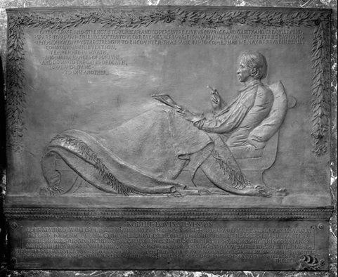 Memorial plaque depicting a reclining figure in relief, draped in a blanket and holding a writing instrument with a book or paper on the lap. The plaque is inscribed with text above and below the figure, framed by a decorative border of leaves.