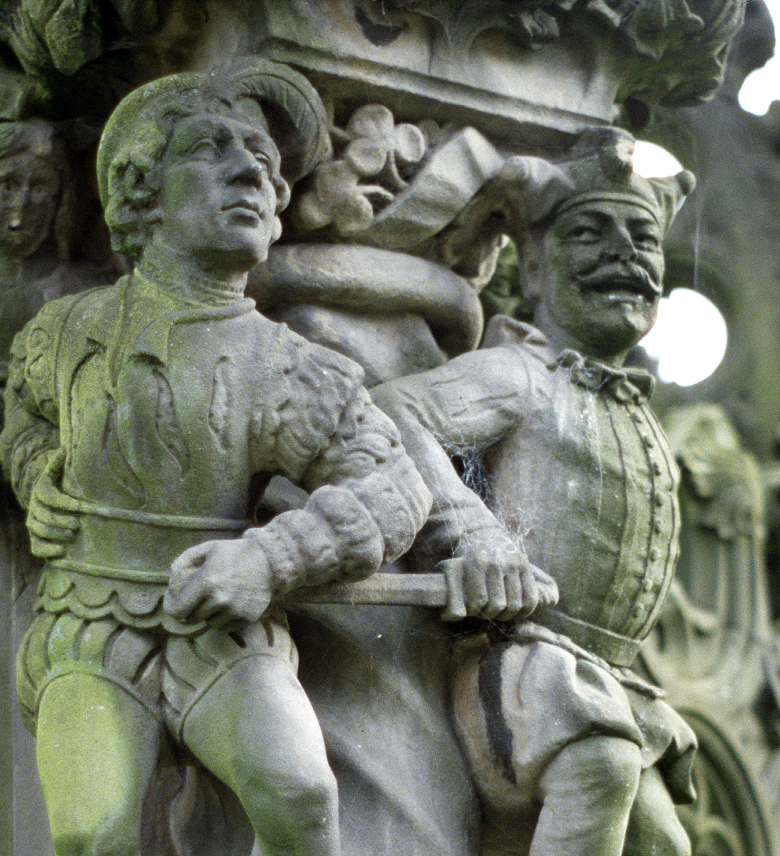 Close-up of an ornate stone carving showing two historical figures in Renaissance-style clothing. On the left, the Duke of Devonshire is depicted in a fitted doublet with puffed sleeves, while on the right George Buchanan, the court fool, is shown thrusting a sword toward the duke. The background features intricate scrollwork and decorative foliage, adding depth and texture to the scene.