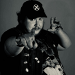 Person wearing a dark cap with a white saltire emblem and a sleeveless jacket decorated with multiple pins. The individual is pointing toward the camera with one hand while the other hand is raised in a dynamic gesture. A graphic T-shirt featuring a monochrome image of a person in sunglasses and a hat is visible beneath the jacket. The photo is in black and white with a plain background.