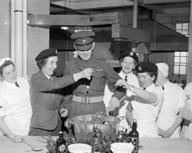 Military personnel and kitchen staff gather around a large mixing bowl at Dreghorn Camp as a Christmas pudding is being prepared. Major D. Fraser stirs the mixture while Major Ravenscroft drops a six‑pence coin into the bowl. Bottles of ingredients and festive greenery sit on the table in front of them.