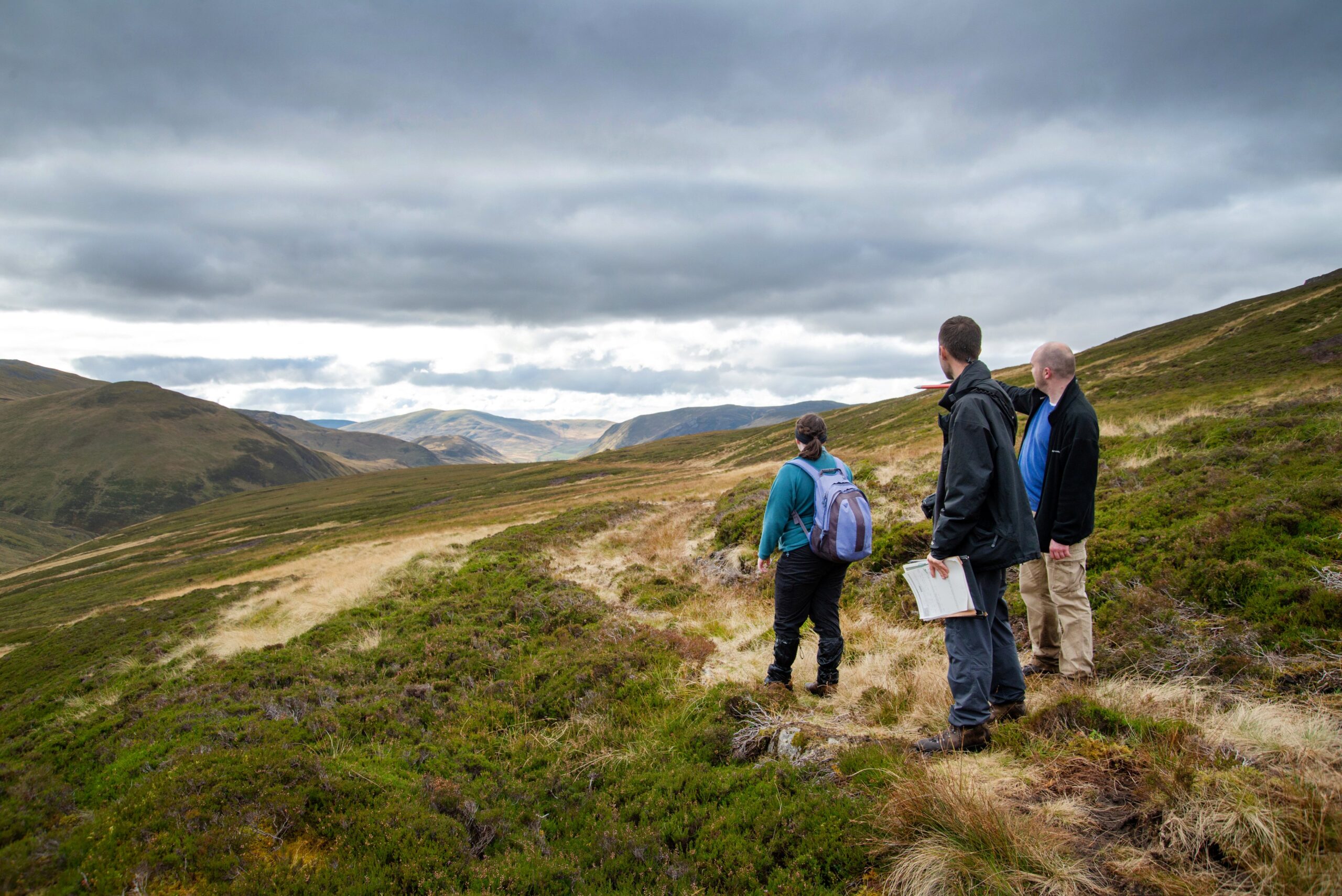 Three people stand on a grassy hillside in a remote, mountainous landscape under a cloudy sky. One person carries a backpack, another holds a map, and they appear to be looking out toward distant hills and valleys.