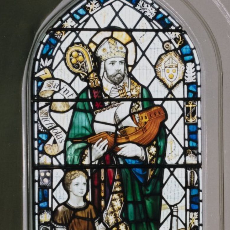 Stained‑glass window depicting Saint Nicholas dressed in ornate religious robes, holding a small model ship. A child stands beside him. The window features decorative borders, heraldic symbols, and a scroll reading “Saint Nicholas.”