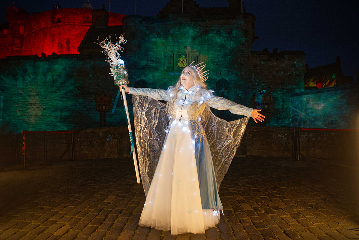 woman in a white gown with a staff and crown standing in front of a castle at night