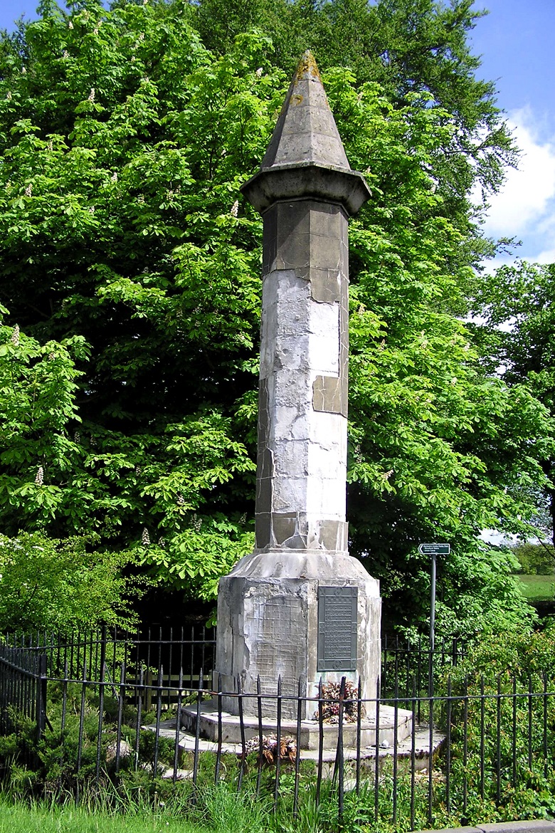 A tall stone memorial column with a pointed top, commemorating the Battle of Falkirk. The monument stands within a small fenced area surrounded by greenery and trees. The base of the column is hexagonal and features a plaque with inscriptions. 