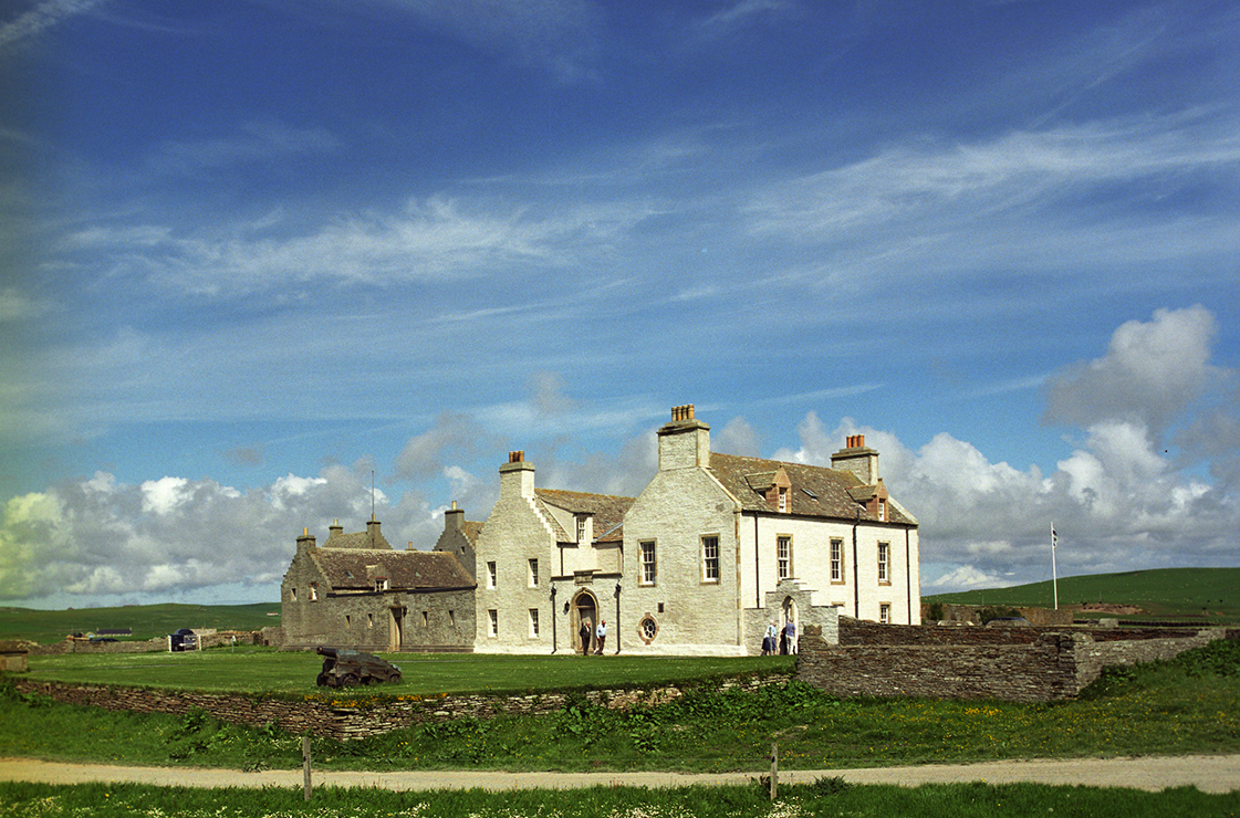 Large stone-built historic house set in open fields. The light-coloured main building has multiple chimneys, tall windows, and an arched entrance. A long, lower stone building stands behind it.