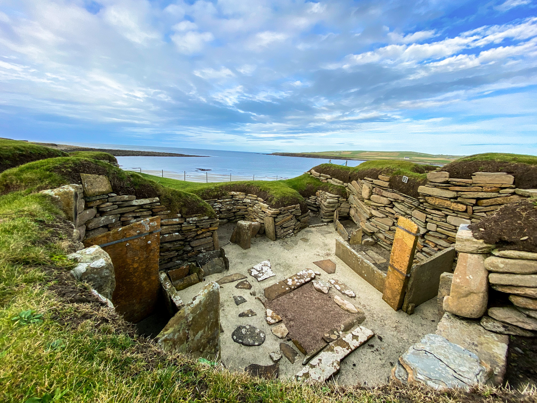 house remains built from stacked stone, forming low rounded walls around a central area. Stone slabs outline former hearths and internal features on a sandy floor. The structure sits in grassy earth mounds overlooking a wide bay