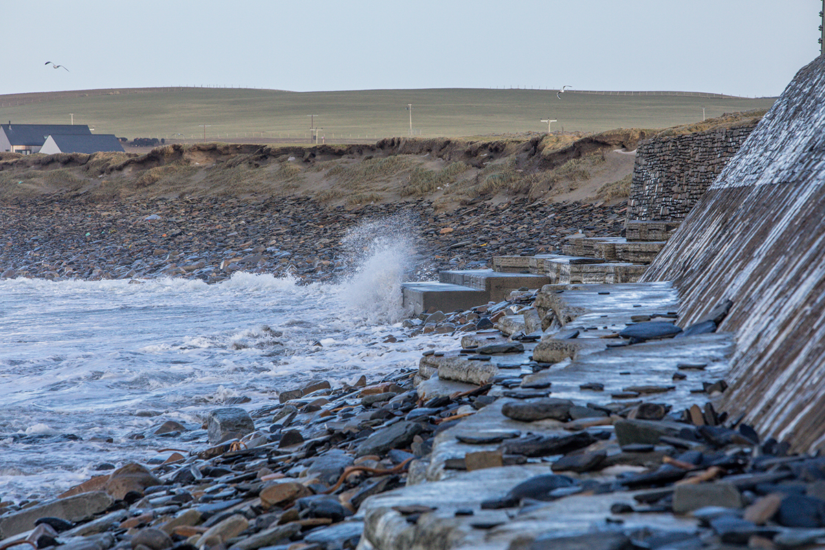 Waves crash against a stone seawall beside a rocky shoreline. Water splashes upward where the tide meets the wall, and wet stones are scattered along the edge. 