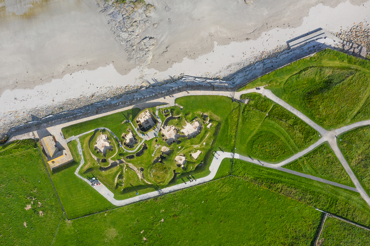 Aerial view of a cluster of low, circular and interconnected stone structures sits near a sandy beach, separated from the shore by a long stone seawall. Paved paths loop around the site and extend across the surrounding grassy landscape.