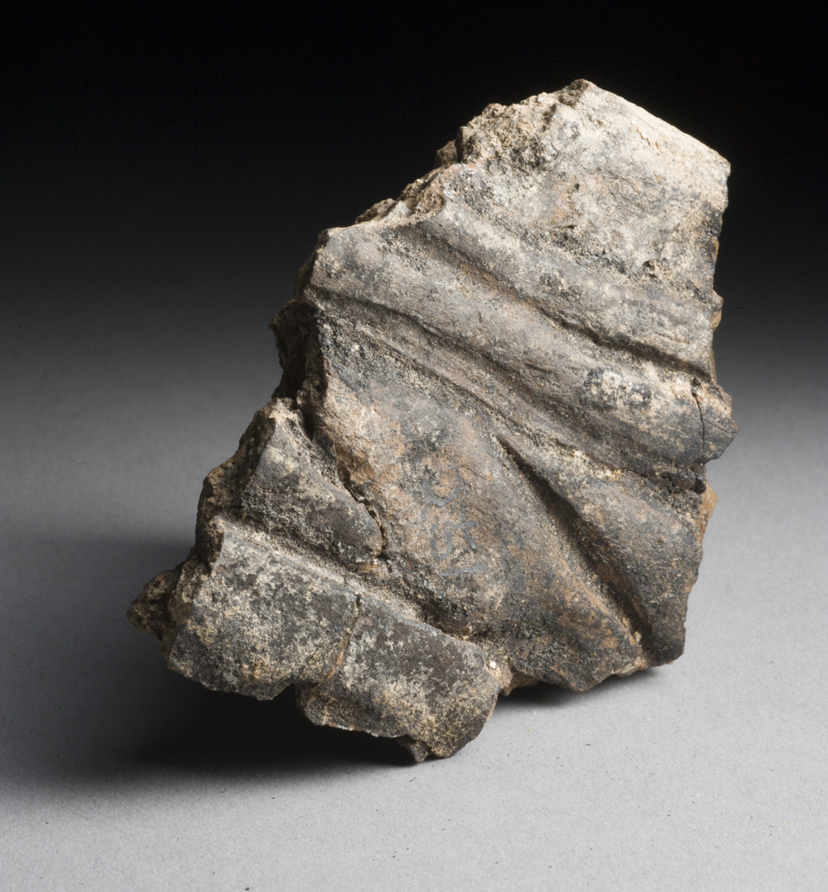 fragment of weathered pottery with raised, diagonal ridged decoration on its surface. The broken edges are rough, and the piece is displayed against a plain, softly lit background.