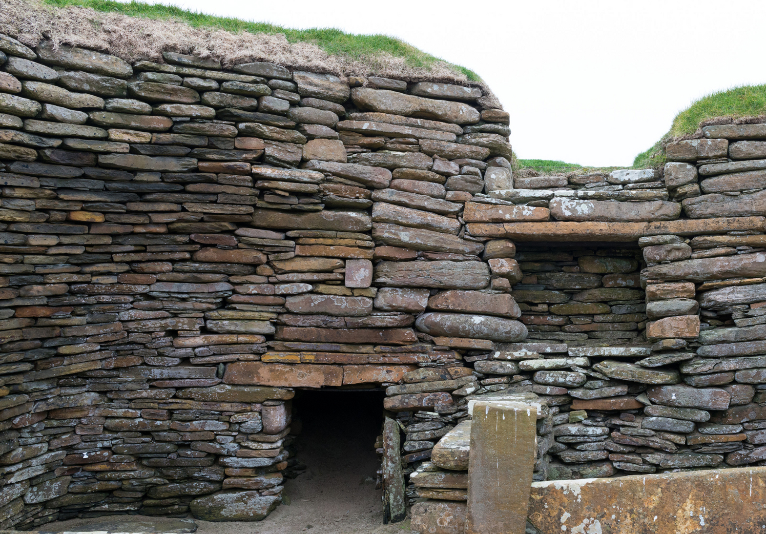 dry‑stone wall made of tightly stacked, flat stones. Near the centre is a low, rectangular doorway leading into a dark interior space. To the right, a small stone shelf or recess is built into the wall. Grass‑covered earth forms the top edge of the structure.