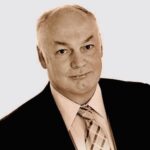  A professionally dressed individual with short, light-colored hair, wearing a dark suit jacket, light dress shirt, and patterned tie, facing the camera with a neutral expression against a plain light background.