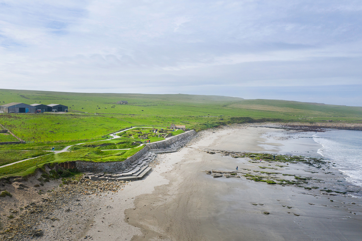 Aerial view of a coastal landscape showing a sandy beach curving beside a stone seawall. Behind the wall, green grassland stretches inland with footpaths leading to a small archaeological site. A few farm buildings stand further back on the open fields