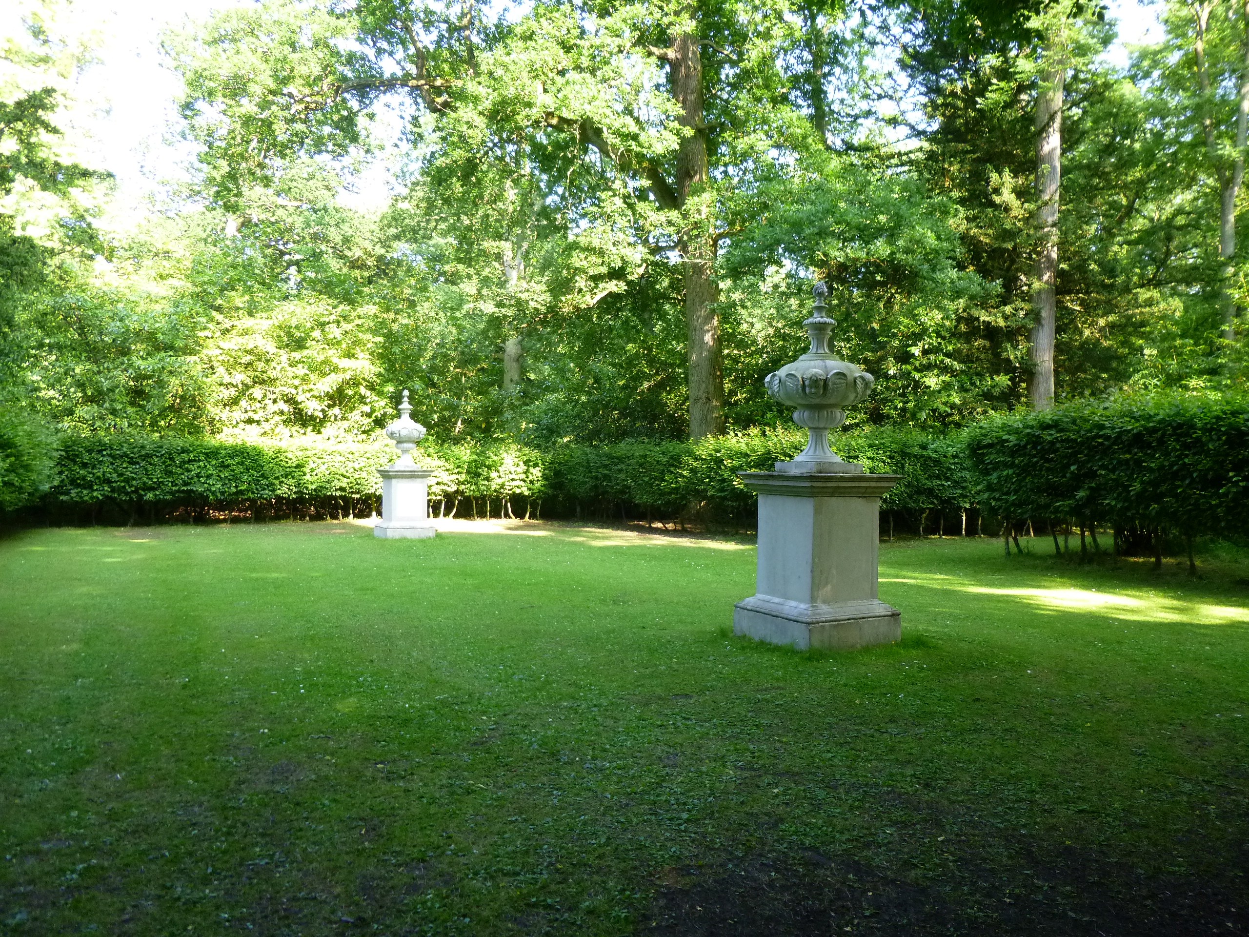 A formal garden clearing with neatly cut grass, containing two stone urns on square pedestals. The urns are aligned one behind the other, surrounded by dense green hedges and tall trees in full leaf.