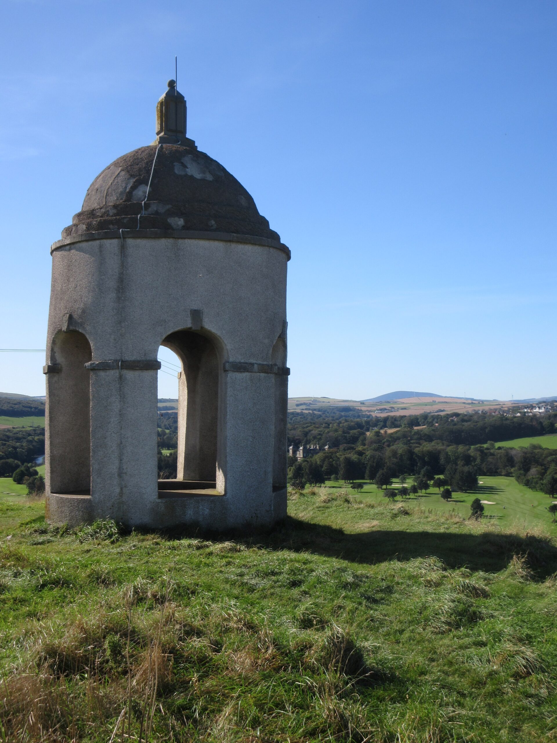 A small domed pavilion with arched openings standing on a grassy hilltop, overlooking a wide valley of rolling green fields under a clear blue sky.