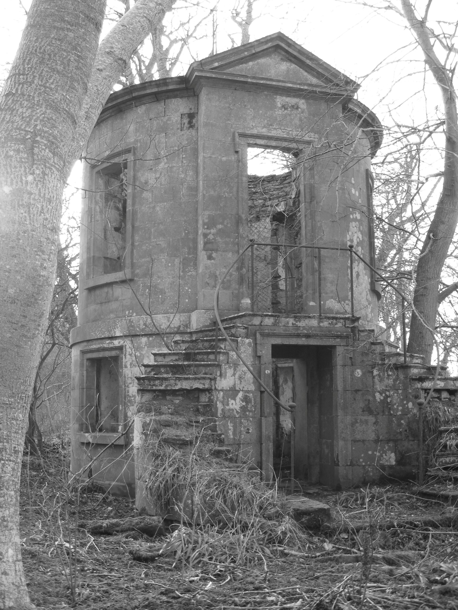 A black‑and‑white photograph of a small, two‑storey stone pavilion in ruins, with an octagonal shape, empty window openings, exterior stairs, and bare trees surrounding the structure.