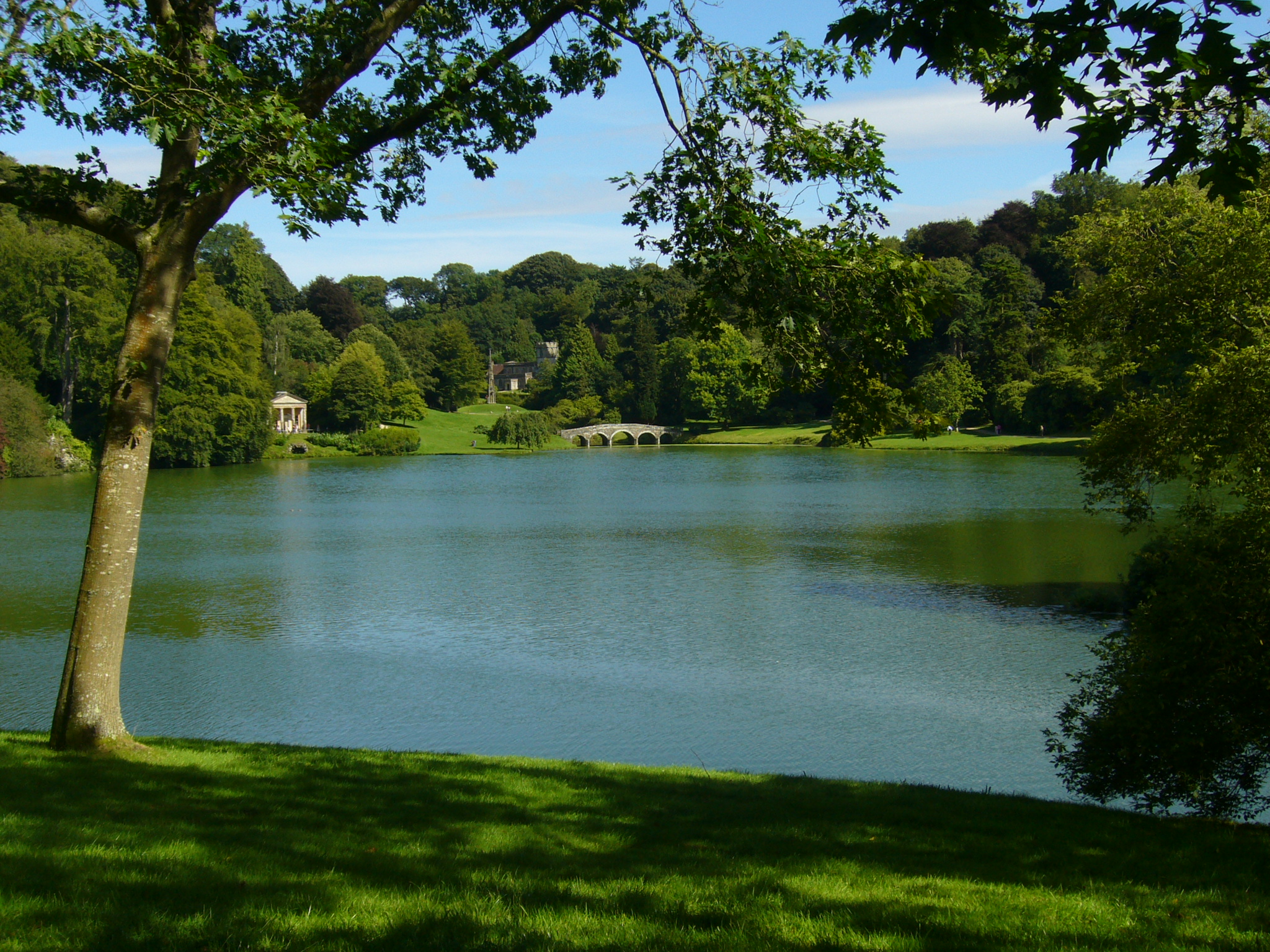A wide lake surrounded by rolling green parkland, with a small arched stone bridge and a classical temple visible among the trees in the distance.