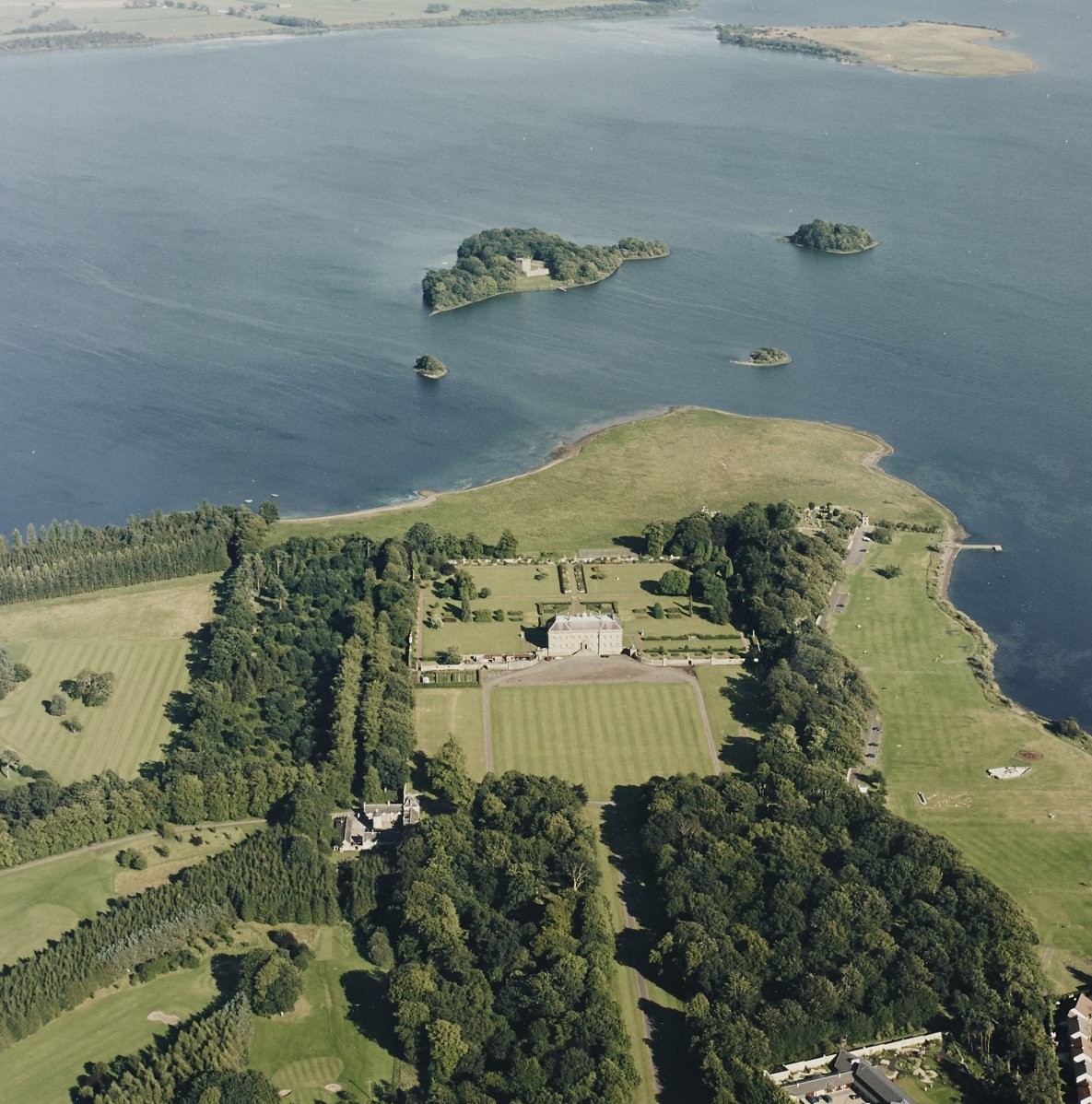 Aerial view of a large historic estate surrounded by formal lawns, tree-lined avenues and woodland, set beside a wide body of water with several small islands.