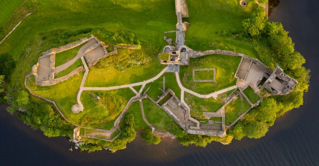 Aerial view of the ruins of Urquhart Castle sitting on a grassy promontory beside Loch Ness, showing the full layout of the remaining stone walls, courtyards, and pathways surrounded by lush greenery and the dark waters of the loch.