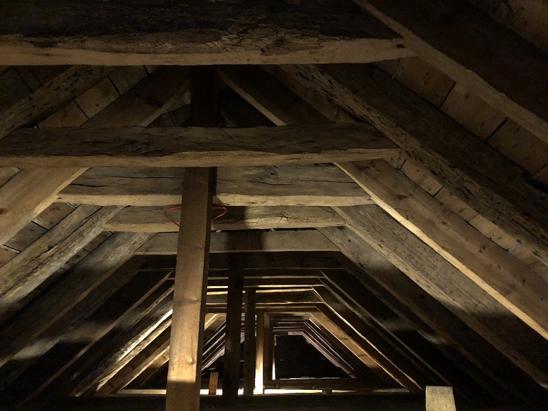 Interior view of an old loft or roof space showing a series of heavy timber beams and rafters. The wooden structure forms repeating triangular frames that recede into the distance, lit by dim, warm light.
