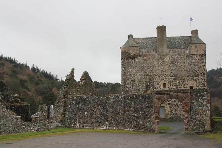 A stone tower house and surrounding ruined walls set against an overcast sky. The structure stands beside wooded hills, with an arched gateway leading into the courtyard.