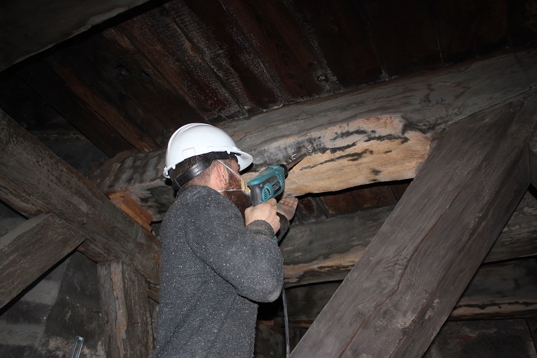Inside a timber roof space, a person wearing protective gear uses a power tool to drill into a large wooden beam. The surrounding beams are dark and aged, suggesting an older structure.