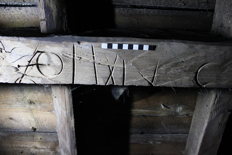 A close-up of a wooden beam inside a roof structure showing carved symbols or markings. A small black‑and‑white measurement scale rests on top of the beam, and cobwebs are visible nearby.