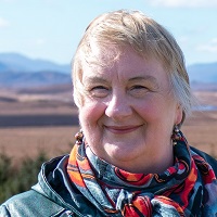A woman wearing a colourful patterned scarf and a dark jacket with short grey hair is standing outdoors in a wide, open landscape. The background shows rolling hills and mountains under a bright blue sky with scattered clouds, with a large expanse of brown moorland stretching into the distance.