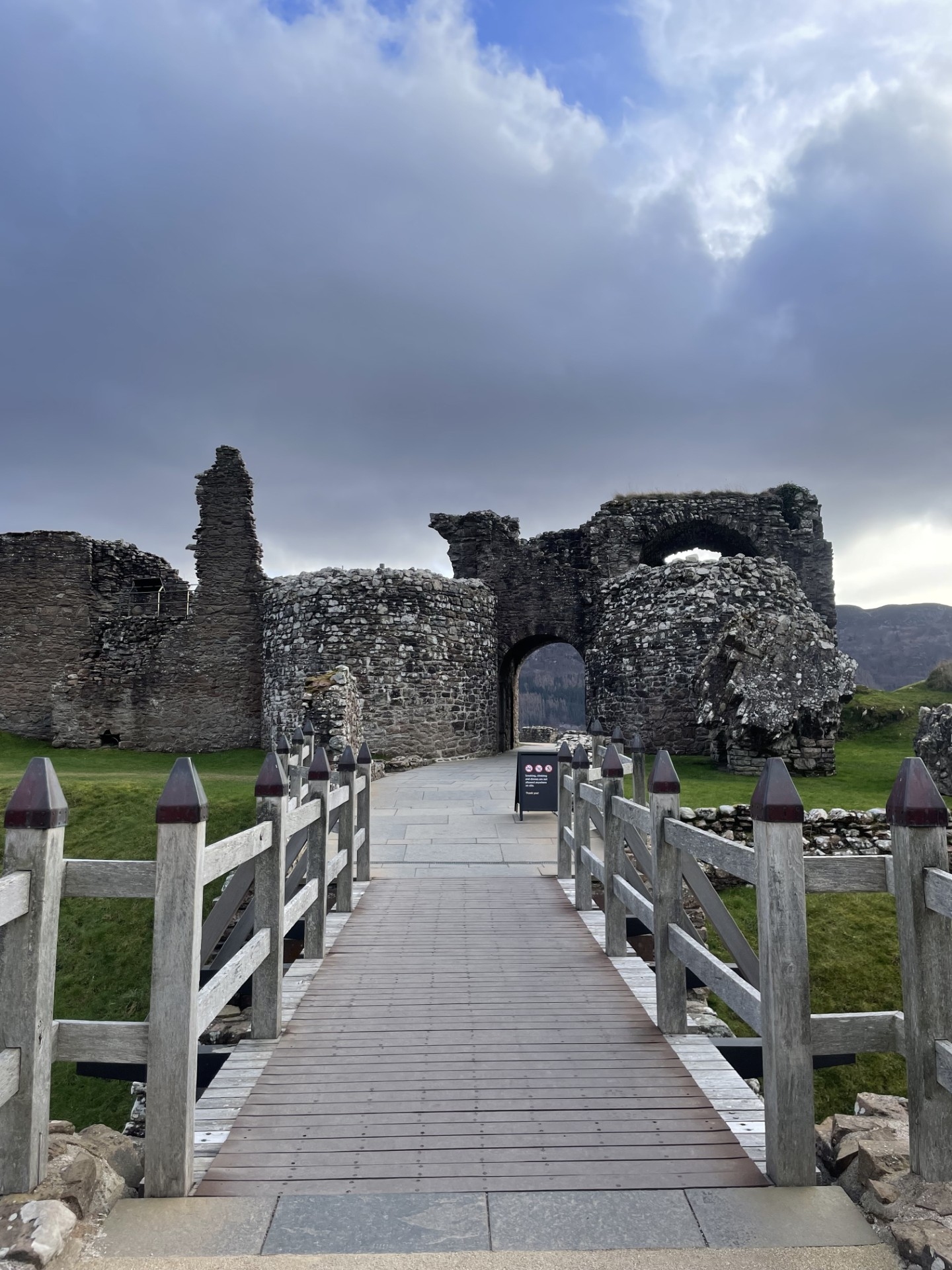 A wooden walkway leading toward a stone archway and the rugged outer walls of Urquhart Castle, set against a cloudy sky.